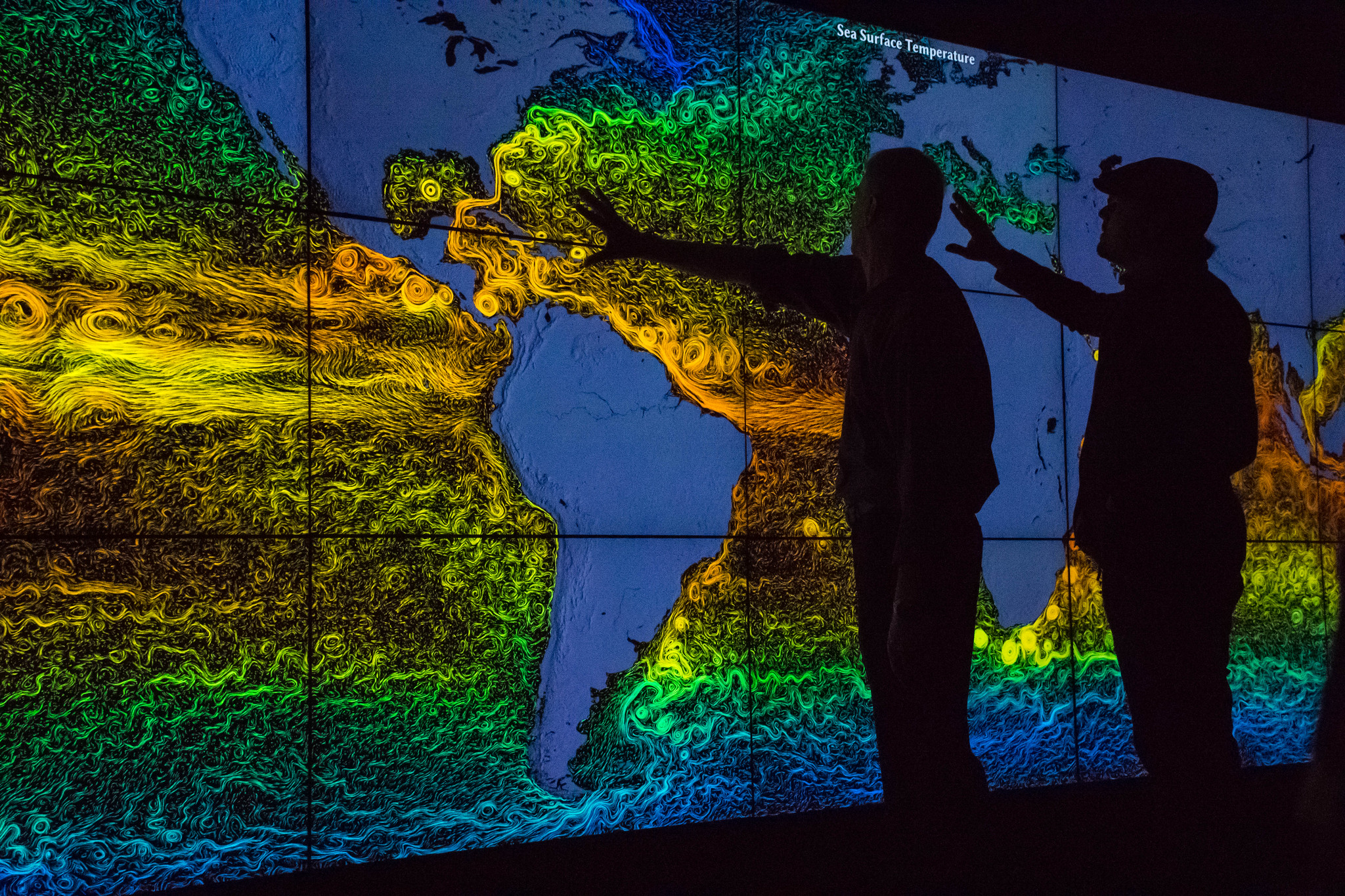 Two people are front lit by a colorful digital map that shows swirly lines on all of the world's oceans.