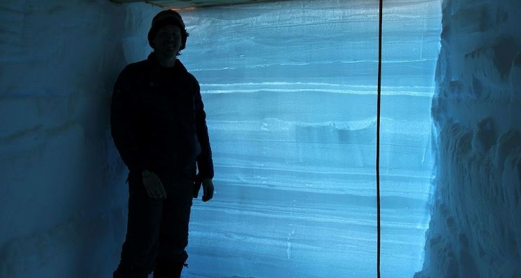 a man standing next to a wall of snow illuminated from the back to show the different layers of snow