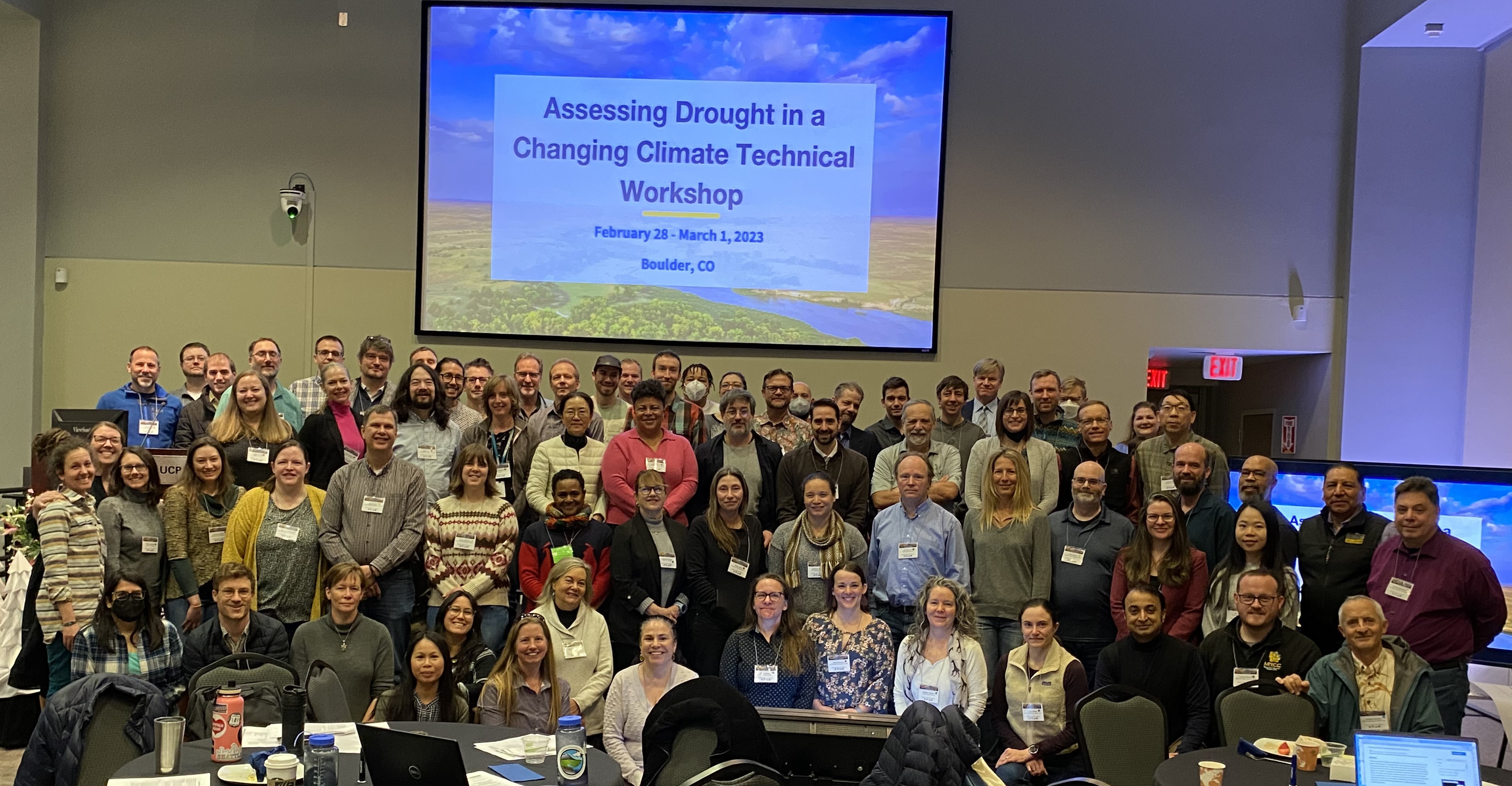 A group of people standing in front of a projector screen that reads Assessing drought in a changing climate technical workshop - February 28-March 1, 2023 - Boulder, CO
