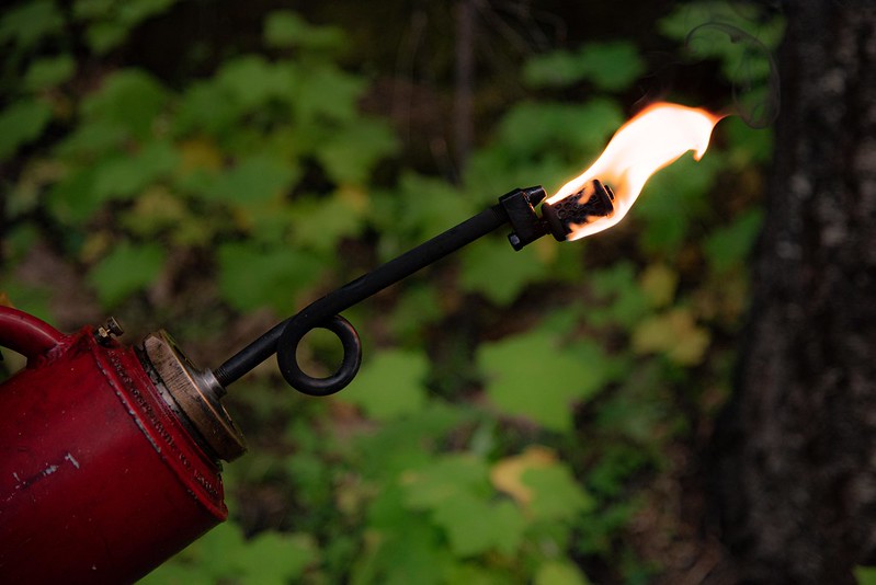A red drip torch with flames emitting from the nozzle. 
