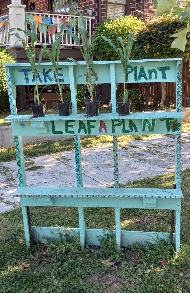 A picture of a home-made two-tiered shelf labeled "take a plant, leaf a plant" with three corn plants on one of the shelves.