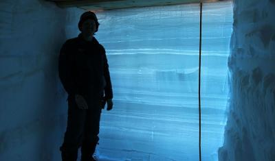A man standing in front of a wall of snow that is backlit, revealing the many different layers of snow.