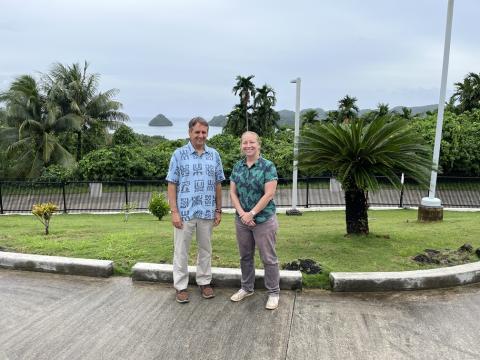 A man and a woman stand smiling on a paved area in front of a manicured grassy area with palm trees, shrubbery, and the ocean in the background.