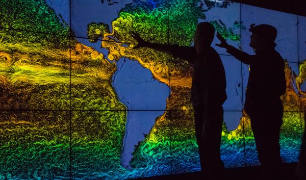 Two people are front lit by a colorful digital map that shows swirly lines on all of the world's oceans.