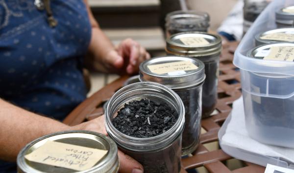 A woman sits behind 5 jars fillled with black biochar, holding one that has the lid off, showing it to the camera