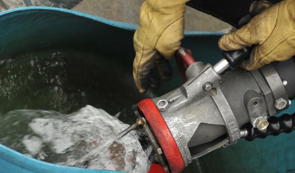 A person wearing tan leather gloves holds a large hose that is filling a turquoise bucket with a mixture of biofuel.