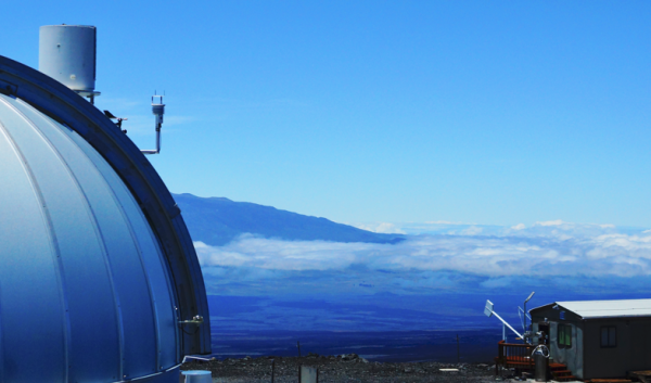 Part of the dome of Mauna Loa observatory looking towards the slopes of a neighboring volcano, with clouds in the distance